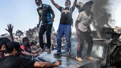 Three young people on top of vandalised vehicle with plume of black smoke amid crowd