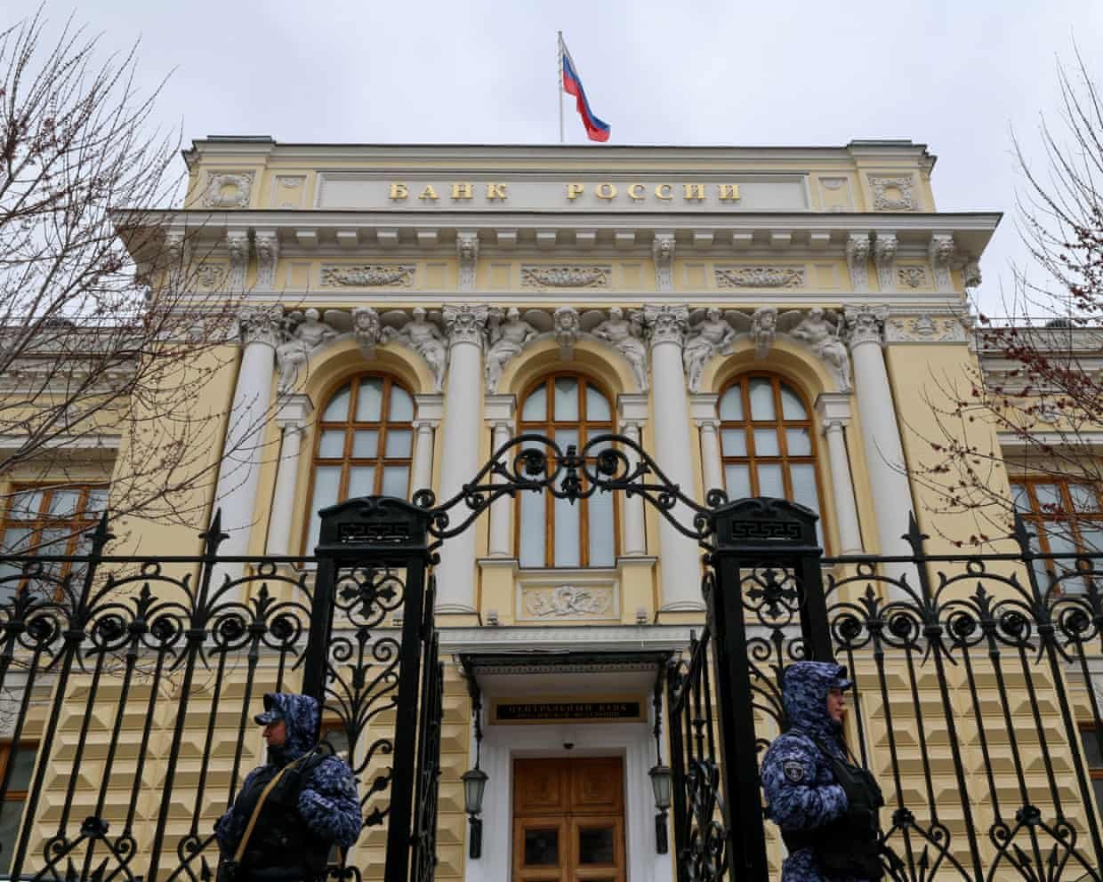 Two guards in blue camouflage uniforms stand outside the Russian Central Bank building with white columns and decorative gates