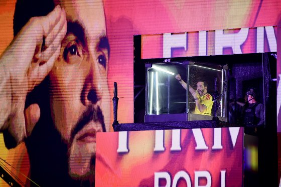 Abelardo de la Espriella raises a fist into the air while speaking into a microphone from inside an elevated, bulletproof glass cube.